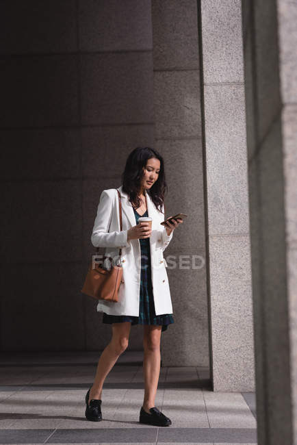 Vista lateral da jovem bela mulher asiática usando telefone celular e tomar café enquanto caminha no corredor — Fotografia de Stock