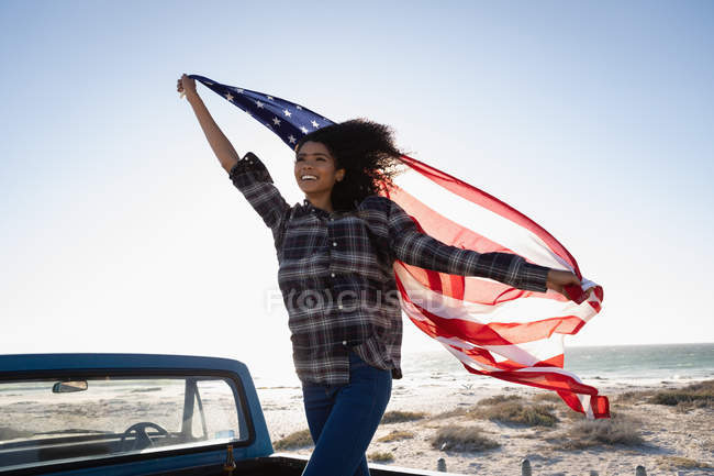 Vista frontal de la hermosa joven afroamericana sosteniendo bandera americana mientras está de pie en el coche en la playa en un día soleado - foto de stock