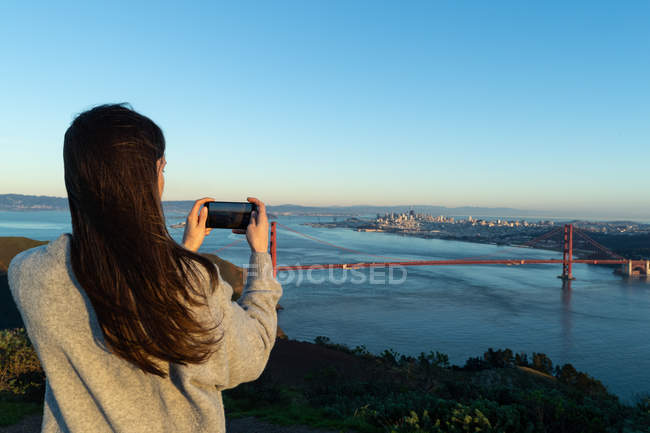 Visão traseira da mulher tirando uma foto de uma ponte em um dia ensolarado — Fotografia de Stock