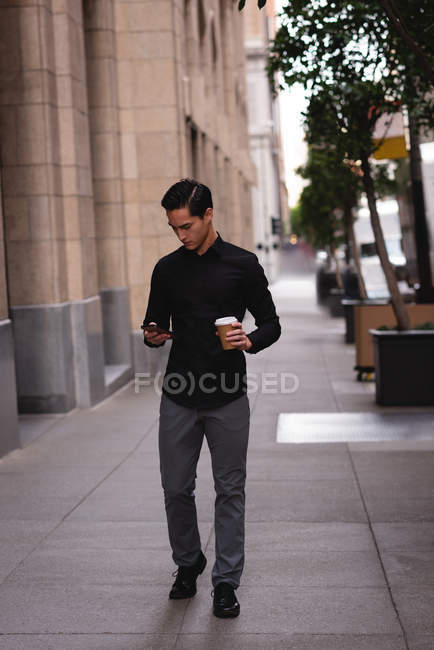 Vista frontal do homem asiático bonito usando telefone celular com café enquanto caminha da rua — Fotografia de Stock