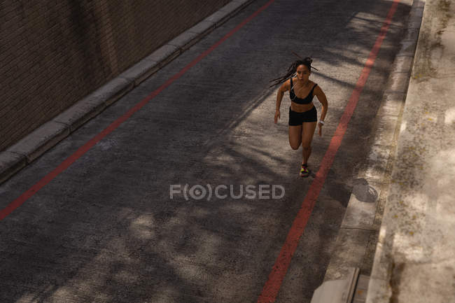 Vue de face de la jeune femme de race mixte écoutant de la musique sur des écouteurs tout en faisant du jogging dans la rue — Photo de stock