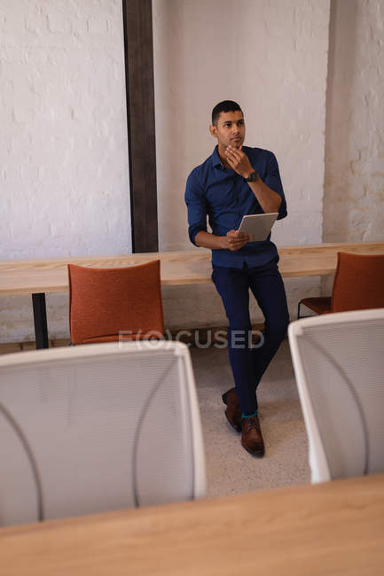 Vista frontal del pensativo hombre de negocios de raza mixta de pie con tableta digital en la oficina moderna - foto de stock