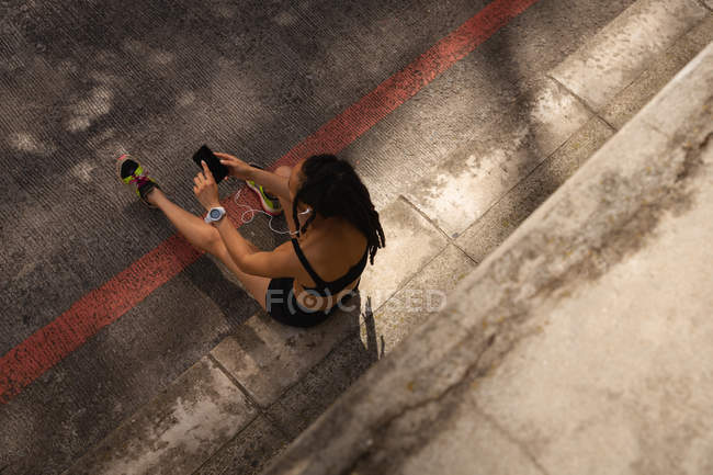 Vista de ángulo alto de ajuste joven mujer de raza mixta escuchando música en el teléfono móvil en la calle - foto de stock