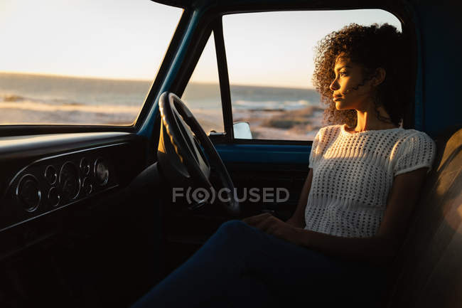 Vista lateral da bela mulher afro-americana pensativa sentada no carro na praia ao pôr do sol — Fotografia de Stock