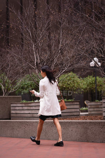 Vista trasera de la mujer asiática usando el teléfono móvil mientras camina en la acera - foto de stock