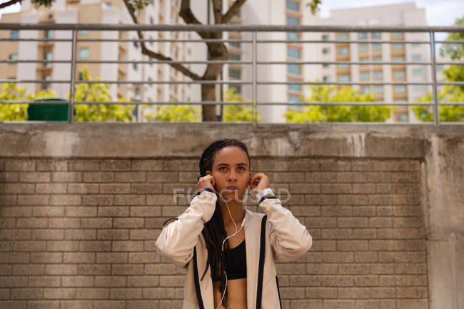 Vue de face de la jeune femme de race mixte écoutant de la musique sur des écouteurs dans la ville — Photo de stock