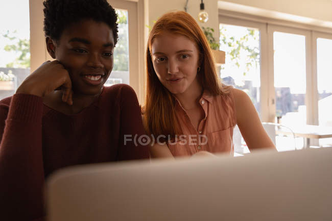 Vista frontal de jovens mestiços amigos do sexo feminino interagindo uns com os outros ao usar laptop no restaurante — Fotografia de Stock