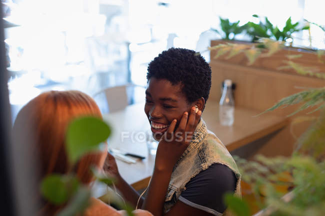 Високий кут зору змішаних раси жіночих друзів прослуховування музики з вухо телефону, сидячи в ресторані — стокове фото