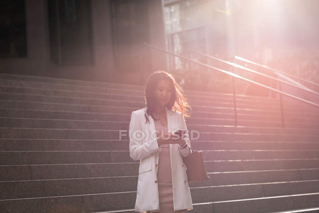 Vista frontal da jovem mulher asiática usando telefone celular enquanto está de pé nas escadas — Fotografia de Stock