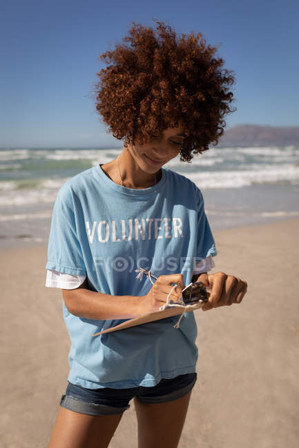 Retrato de jovem mestiça escrevendo em prancheta na praia em um dia ensolarado — Fotografia de Stock