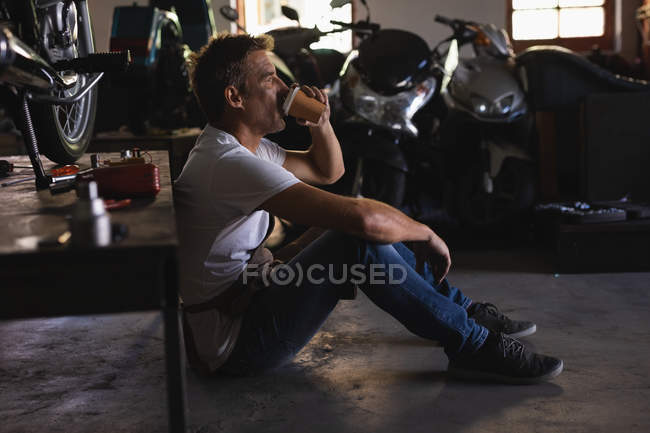 Seitenansicht eines kaukasischen Fahrradmechanikers, der auf dem Boden sitzt, während er in der Garage Kaffee trinkt — Stockfoto