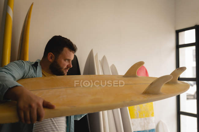 Vista lateral del hombre caucásico revisando minuciosamente la tabla de surf en un taller - foto de stock