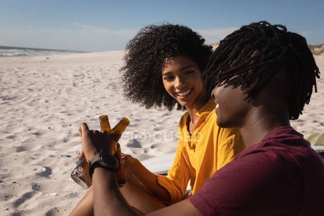 Vista lateral de una pareja afroamericana brindando botella de cerveza en la playa en un día soleado - foto de stock