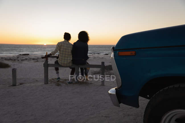 Vue arrière du couple romantique assis sur la plage au coucher du soleil — Photo de stock