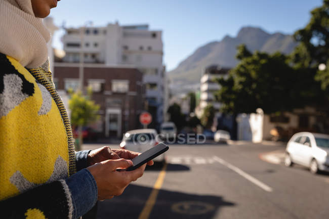 Seitenansicht einer Frau mit gemischter Rasse, die an einem sonnigen Tag mitten auf der Straße steht und ihr Handy benutzt — Stockfoto