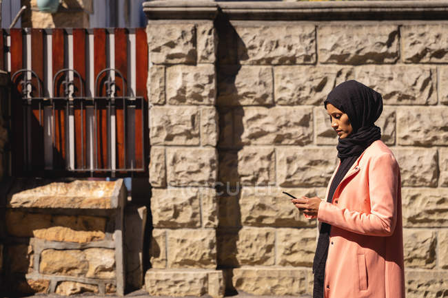 Vista lateral de la mujer de raza mixta usando el teléfono móvil mientras está de pie frente a la pared en la calle - foto de stock