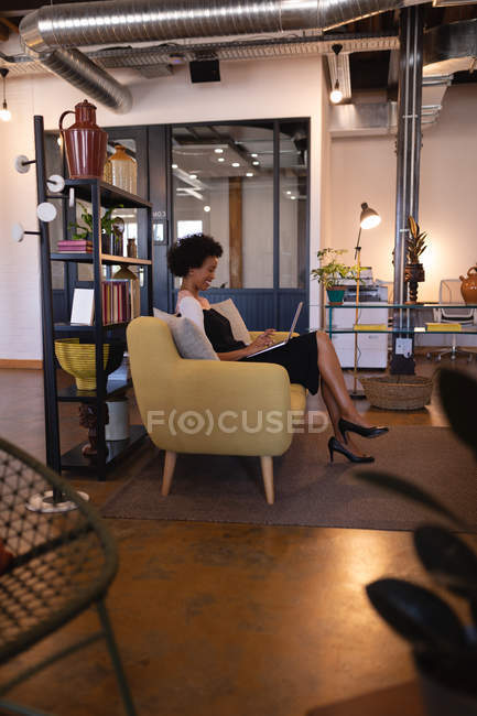 Vue latérale de la belle femme d'affaires mixte utilisant un ordinateur portable au bureau alors qu'elle est assise sur un canapé jaune moderne contre l'ameublement — Photo de stock