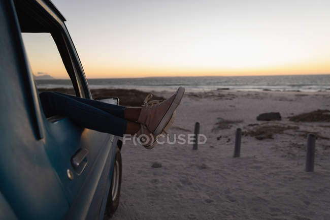 Partie basse de la femme se détendre avec les pieds dans une voiture sur la plage au coucher du soleil — Photo de stock