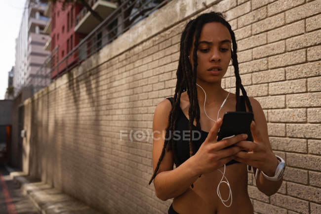 Vista frontal de la joven mujer de raza mixta usando teléfono móvil en la calle de la ciudad - foto de stock