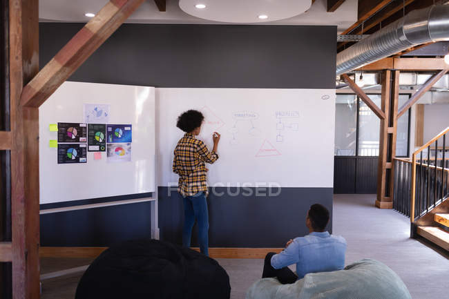 Rückansicht einer jungen Geschäftsfrau mit gemischter Rasse, die auf einer weißen Tafel schreibt, während seine Kollegen sie in einem Sitzsäckchenstuhl in einem modernen Büro betrachten — Stockfoto