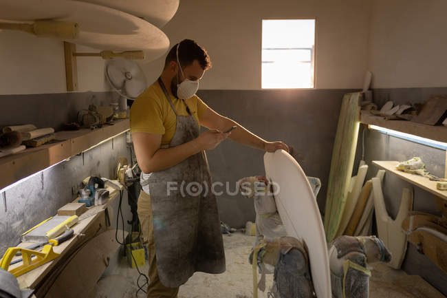Side view of Caucasian man with mouth protection mask using mobile phone while cleaning surfboard in a workshop — Stock Photo