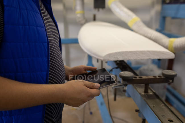 Sección media del hombre de pie y el uso de tableta digital en el taller con una tabla de surf en el fondo - foto de stock