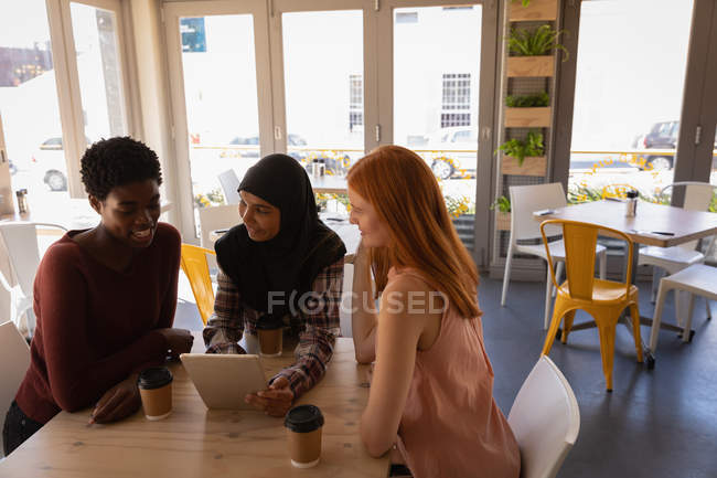 Вид збоку на молоду змішану расу друзі-жінки взаємодіють один з одним, використовуючи цифровий планшет у кафе — стокове фото