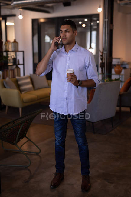 Вид спереду красивий молодий бізнесмен змішаної раси стоїть і розмовляє на мобільному телефоні в сучасному офісі, поки він тримає чашку кави — стокове фото