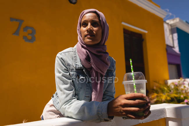 Vista de ángulo bajo de la mujer de raza mixta reflexiva inclinándose mientras tiene jugo en un día soleado en la calle - foto de stock