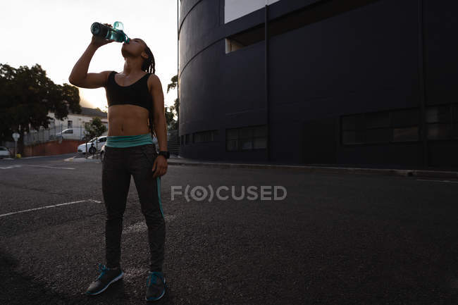 Vue de face de la jeune femme fatiguée de race mixte buvant de l'eau dans la rue — Photo de stock