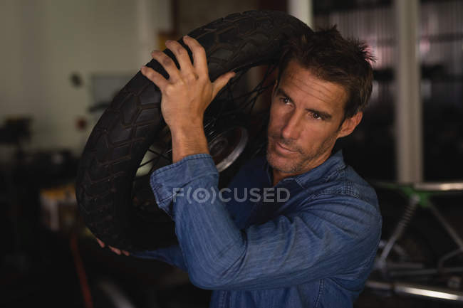 Vista frontal da bicicleta masculina caucasiana mecânico segurando pneu enquanto em pé na garagem — Fotografia de Stock