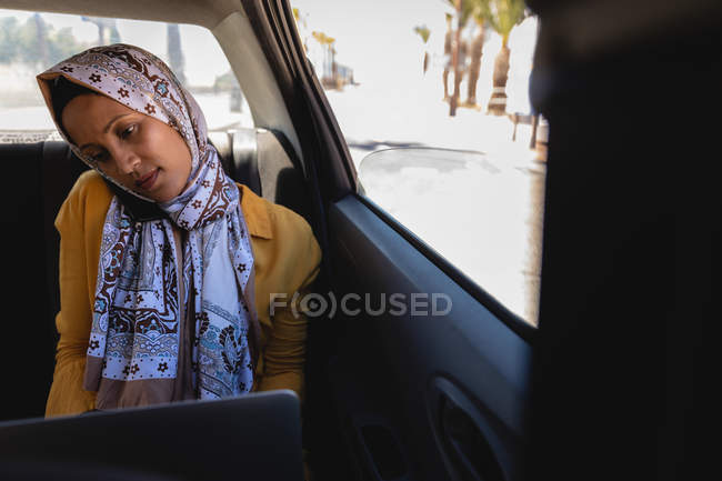 Вид спереду змішаної раси жінка розмовляє на мобільному телефоні, використовуючи ноутбук в машині в сонячний день — стокове фото