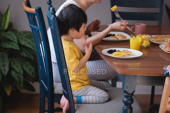 Vista lateral de la madre asiática y el hijo disfrutando del desayuno en la mesa de comedor en la cocina en casa - foto de stock