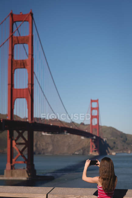 Vista trasera de mujer caucásica tomando foto con teléfono móvil de puente colgante - foto de stock