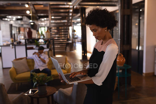 Vista lateral de la hermosa mujer de negocios mixta de pie y el uso de la computadora portátil en la oficina moderna, mientras que su colega con auriculares de realidad virtual en el sofá - foto de stock
