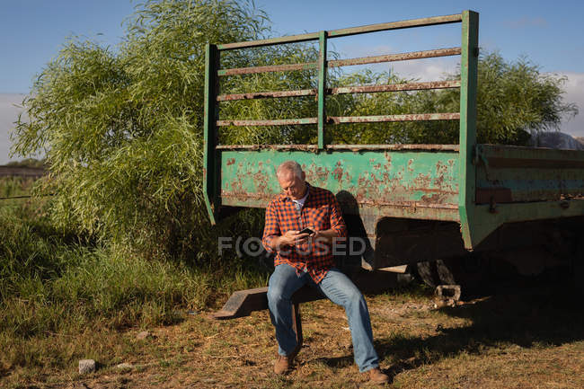 Vue de face du vieil homme caucasien utilisant son téléphone portable assis sur une remorque verte par une journée ensoleillée — Photo de stock