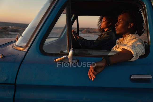 Vista lateral do casal romântico afro-americano dirigindo um carro na praia ao pôr do sol — Fotografia de Stock