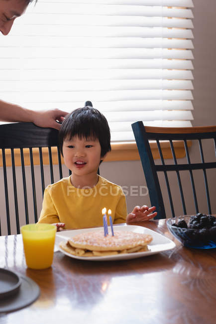 Vista frontal del hijo asiático disfrutando de su cumpleaños frente a panqueques con velas en la mesa de comedor en la cocina en casa - foto de stock