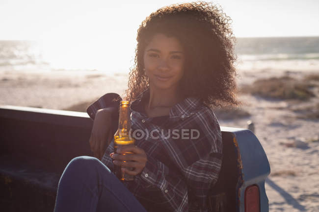 Vista frontal da bela jovem afro-americana sentada no carro enquanto bebe cerveja na praia em um dia ensolarado — Fotografia de Stock