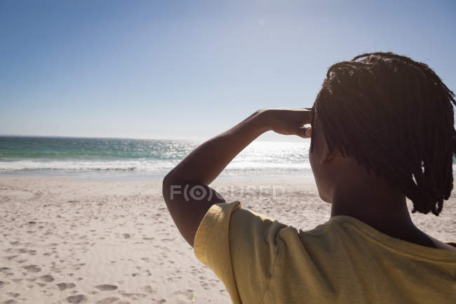Vista posteriore del giovane uomo afroamericano guardando con occhio schermante la spiaggia nella giornata di sole — Foto stock