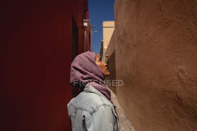 Вид сбоку на вдумчивую расовую женщину, смотрящую вверх, стоя в переулке в солнечный день — стоковое фото