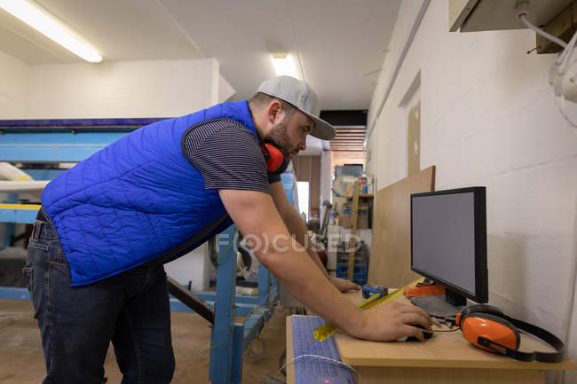 Seitenansicht eines konzentrierten kaukasischen Mannes mit Ohrenschützern um den Hals, der in der Werkstatt am Computer arbeitet — Stockfoto