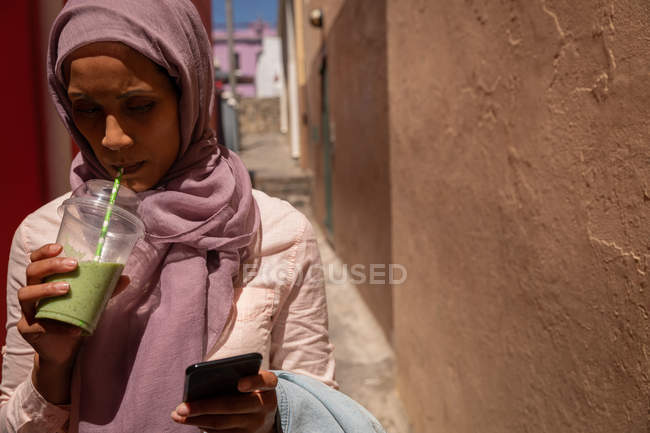 Vue de face de belle femme mixte utilisant un téléphone portable tout en buvant un smoothie par une journée ensoleillée — Photo de stock