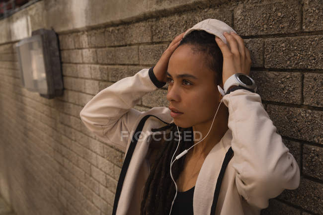 Vista laterale di una bella giovane donna di razza mista che ascolta musica con le cuffie in piedi contro un muro in strada — Foto stock