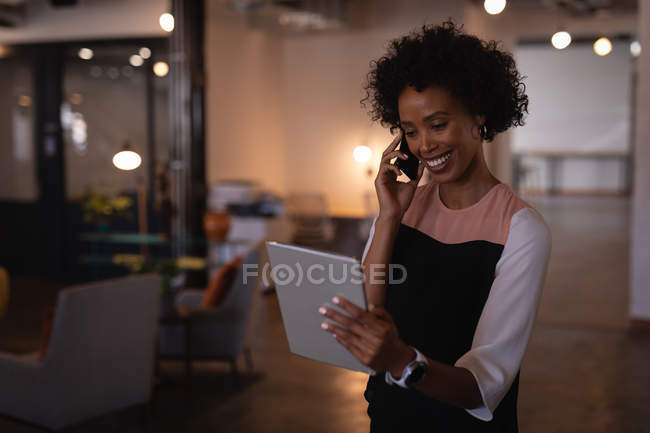 Vue de face de belle jeune femme d'affaires mixte parlant sur téléphone mobile tout en regardant tablette numérique dans le bureau — Photo de stock