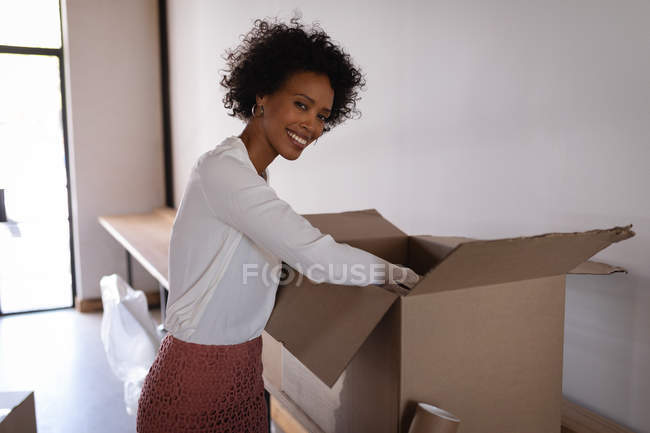 Retrato de feliz mulher de negócios de raça mista desempacotar caixa de papelão no escritório moderno. Ela está olhando e sorrindo para a câmera — Fotografia de Stock