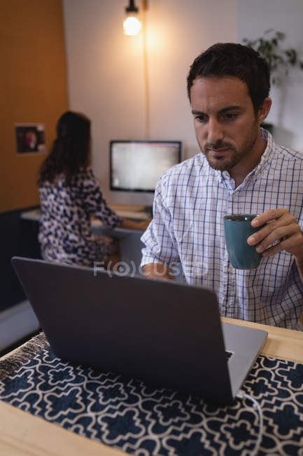 Vista frontal do executivo masculino caucasiano segurando café enquanto trabalhava em laptop na mesa no escritório — Fotografia de Stock