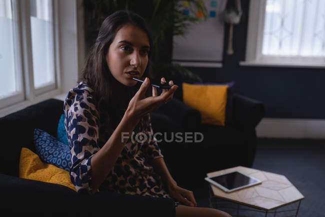 Vista lateral del joven ejecutivo femenino de raza mixta hablando por teléfono móvil mientras está sentado en un sofá en el vestíbulo de la oficina - foto de stock