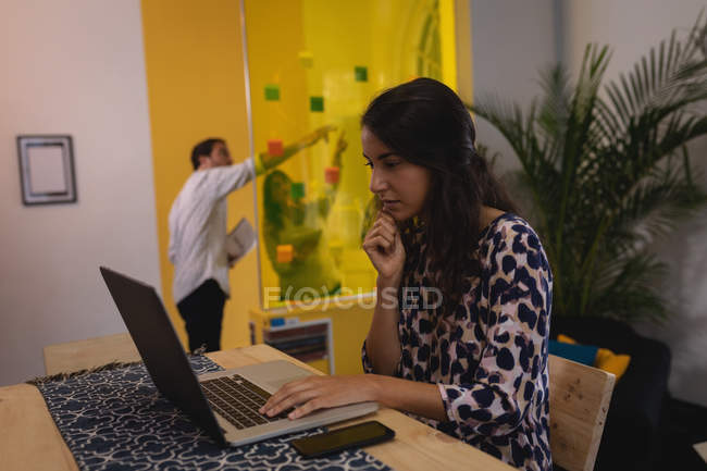 Vue latérale d'un exécutif féminin métis réfléchi travaillant sur un ordinateur portable au bureau pendant que les gens d'affaires discutent sur des notes collantes sur un mur de verre — Photo de stock