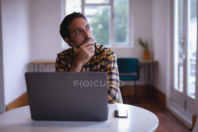 Vista frontal do executivo masculino caucasiano pensativo olhando para longe ao usar o laptop no escritório — Fotografia de Stock